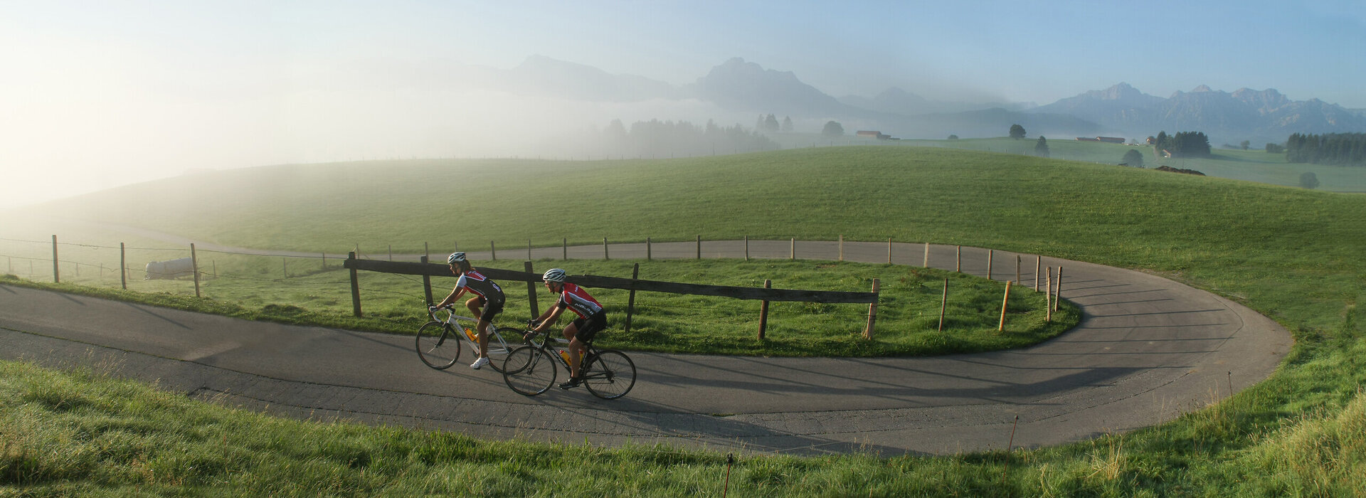 Das Bild zeigt zwei Rennradfahrer in Trikots und Radlerhosen, die einen Hügel emporfahren. Der Morgennebel lichtet sich langsam. Im Hintergrund erheben sich die Berge.  