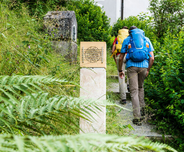 Das Bild zeigt einen Mann und eine Frau in Wanderkleidung mit Rucksäcken. Sie wandern einen Hang empor auf einem Kiesweg mit Stufen. Im Vordergrund steht eine steinerne Säule. Um sie herum wachsen Büsche, Bäume und Gräser.