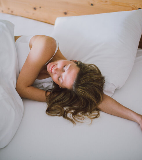 Das Bild zeigt eine Frau, die in einem Bett liegt, das weiß bezogen ist. Ihr Kopf liegt auf ihrem linken Arm. Sie hat die Augen geschlossen und wirkt entspannt.