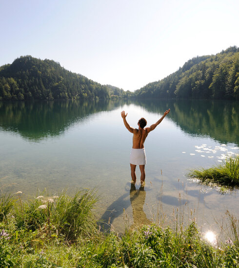 Das Bild zeigt einen Mann, der sich nur ein weißes Handtuch um die Hüften gebunden hat. Er steht im knöcheltiefen Wasser eines Sees und streckt die Arme nach oben.